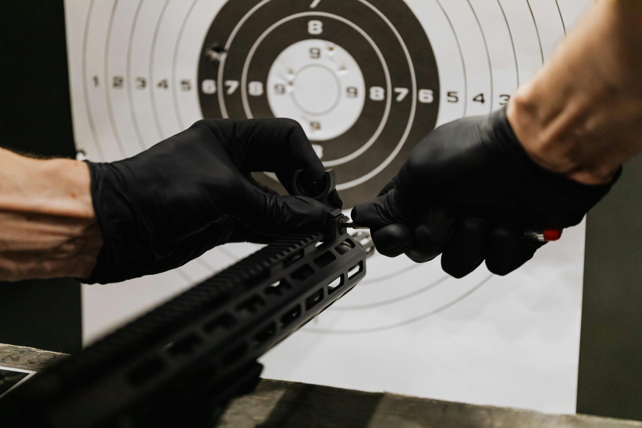 Gloved hands adjusting a rifle's iron sight with a target in the background at a shooting range.